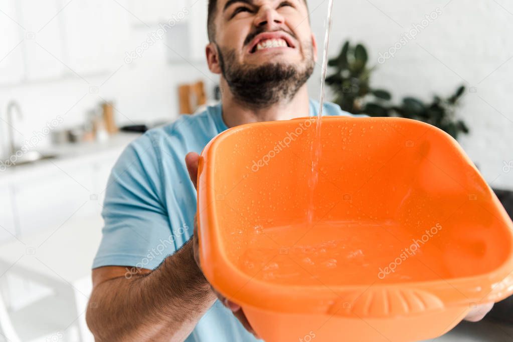 selective focus of displeased bearded man holding plastic wash bowl near pouring water 