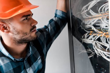 handsome technician in safety helmet looking at wires and cables  clipart