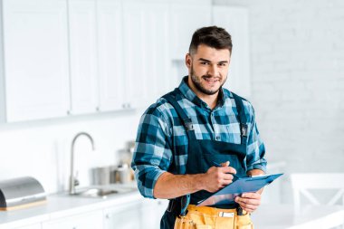 happy bearded workman holding clipboard and looking at camera in kitchen  clipart