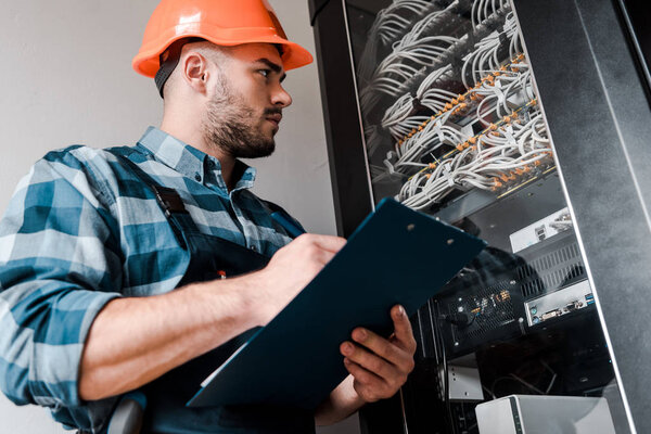 low angle view of bearded man in safety helmet holding clipboard and looking at wires and cables 