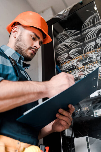 low angle view of bearded workman in safety helmet holding clipboard near wires and cables 