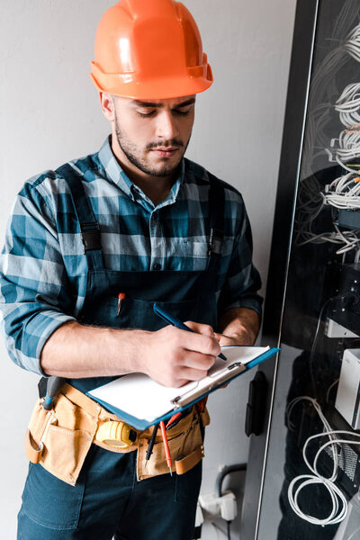handsome technician holding clipboard while writing near wires and cables 