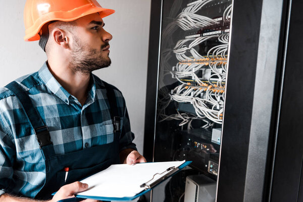 technician in safety helmet holding clipboard while looking at wires and cables 