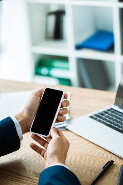  masada dizüstü bilgisayarın yanında boş ekranlı akıllı telefonu tutan adamın kırpılmış görünümü 