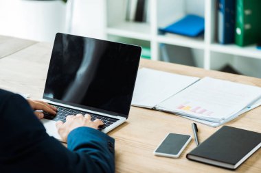 boş ekranlı akıllı telefonun yanındaki dizüstü bilgisayarda yazan işadamının kırpılmış görünümü