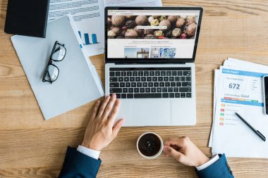 KYIV, UKRAINE - JULY 8, 2019: top view of businessman holding cup near laptop with depositphotos website on screen  clipart