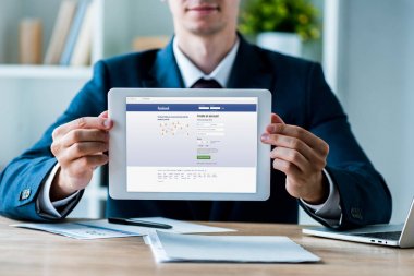 Kiev, Ukrayna - 8 Temmuz 2019: ofiste dizüstü bilgisayarın yanında facebook uygulaması yla dijital tablet tutan adamın seçici odağı 