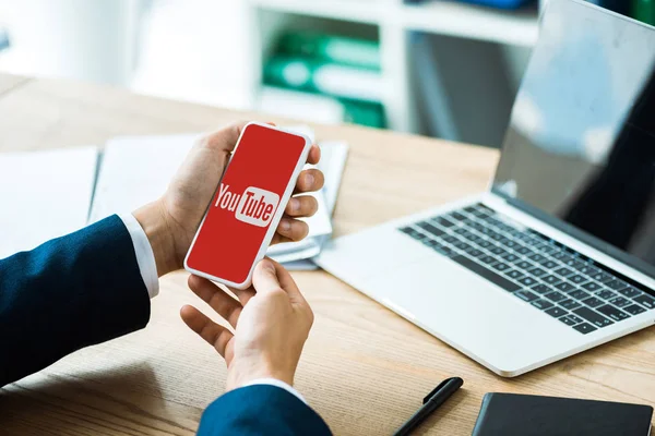 Kiev, Ukrayna - 8 Temmuz 2019: masada dizüstü bilgisayar ve kalem yakınında youtube uygulaması ile akıllı telefon tutan adam kırpılmış görünümü 
