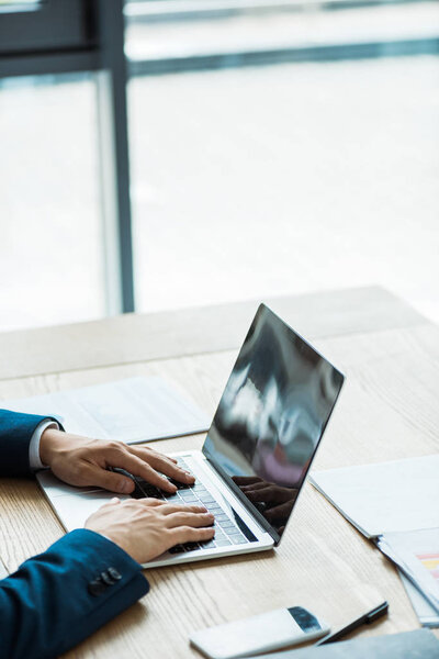 cropped view of man typing on laptop near smartphone with blank screen