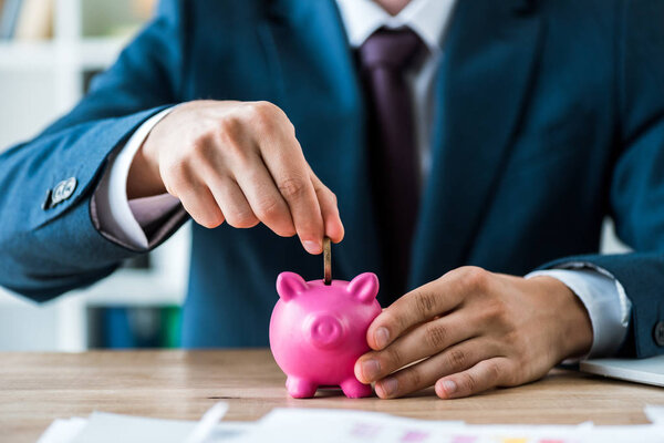 selective focus of man putting metallic coin into piggy bank near laptop