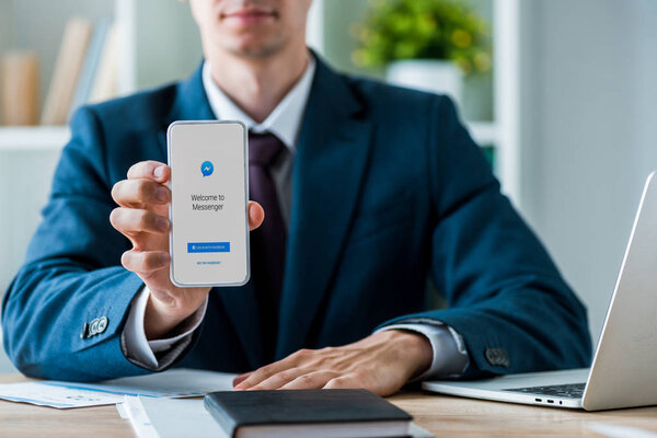 KYIV, UKRAINE - JULY 8, 2019: selective focus of man holding smartphone with messenger app on screen near laptop in office 