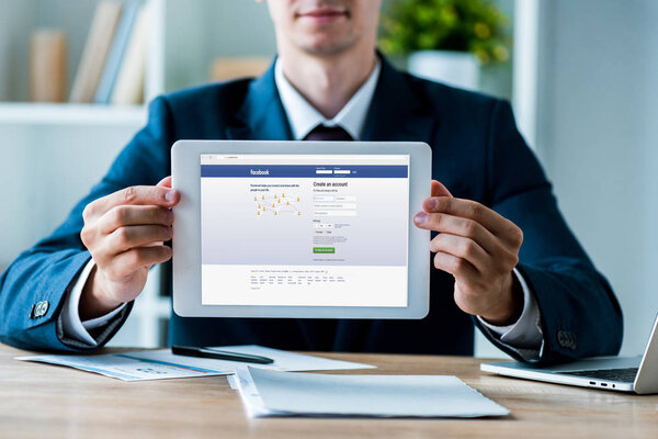 KYIV, UKRAINE - JULY 8, 2019: selective focus of man holding digital tablet with facebook app on screen near laptop in office 