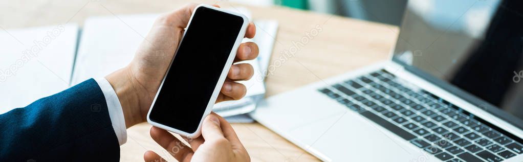 panoramic shot of man holding smartphone with blank screen near laptop on table 
