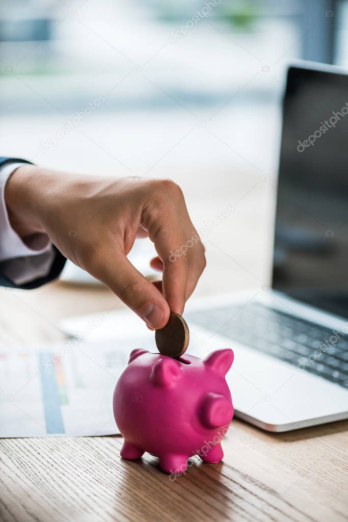 cropped view of businessman putting coin into piggy bank near laptop