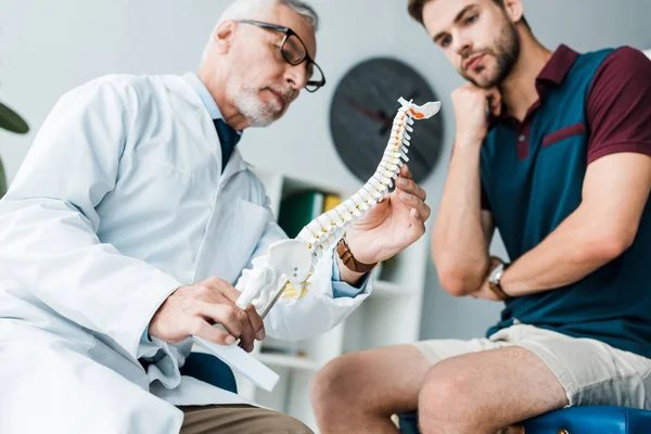 hastanede hastanın yanında omurga modeli tutan sakallı doktor düşük açı görünümü 
