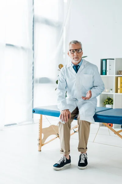 Bebaarde Doctor Glazen Witte Vacht Zittend Massagetafel Gebaren Kliniek — Stockfoto