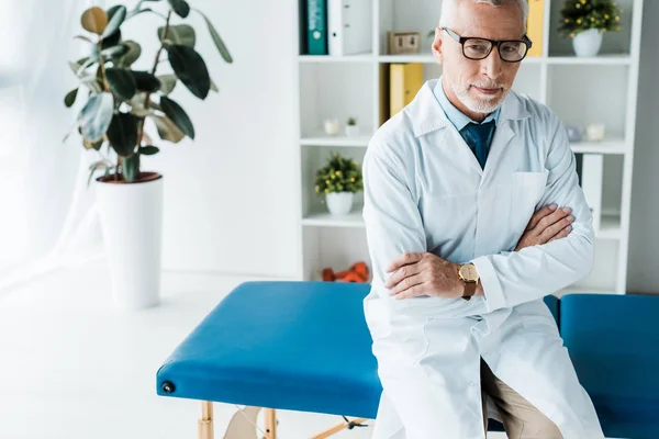 Šťastný Vousatý Doktor Brýlích Bílém Plášti Sedící Masážním Stolku Zkřížené — Stock fotografie