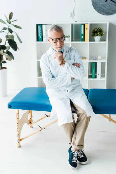Στοχαστικός Γιατρός Γυαλιά Και Λευκό Παλτό Κάθεται Στο Τραπέζι Μασάζ — Φωτογραφία Αρχείου