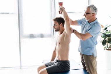 chiropractor standing near man exercising with dumbbell in clinic  clipart