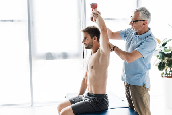 chiropractor standing near man exercising with dumbbell in clinic 