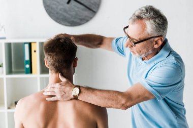 klinikte oturan gömleksiz hastanın boynuna dokunan gözlüklü sakallı doktor 