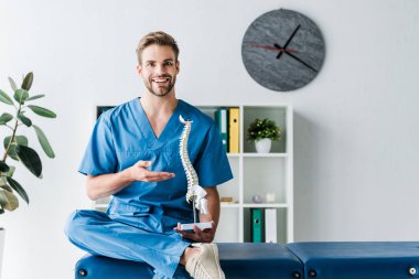 cheerful doctor looking at camera while gesturing and holding spine model in clinic  clipart