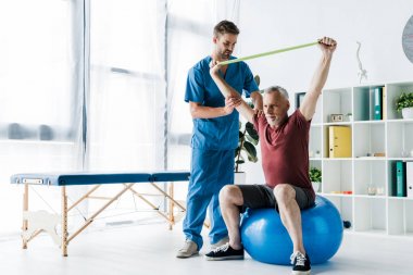 fitness topu otururken direnç bandı ile egzersiz orta yaşlı adam yakınında duran doktor 