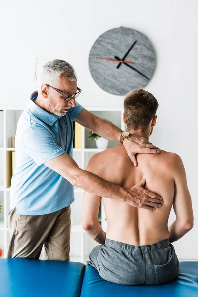 Bearded Doctor Glasses Touching Back Shirtless Patient Sitting Clinic — Stock Photo, Image