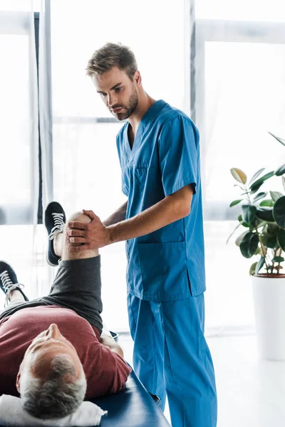 Knappe Arts Aanraken Been Van Volwassen Patiënt Uitoefenen Massagetafel — Stockfoto