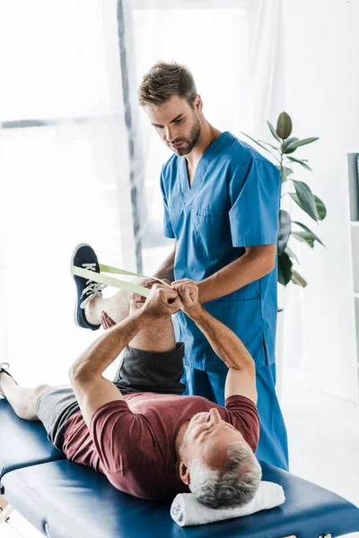 Guapo Doctor Sosteniendo Pierna Maduro Paciente Ejercitando Con Elásticos — Foto de Stock