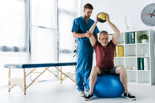 handsome doctor standing near mature man exercising on fitness ball 