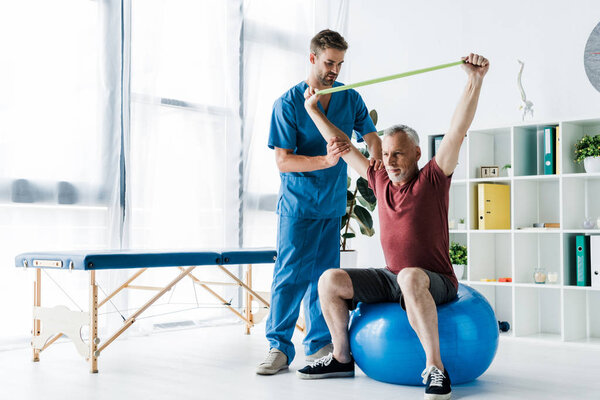 doctor standing near middle aged man exercising with resistance band while sitting on fitness ball 