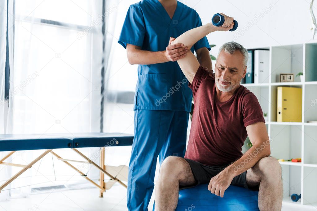 cropped view of doctor standing near middle aged man exercising on fitness ball with dumbbell 