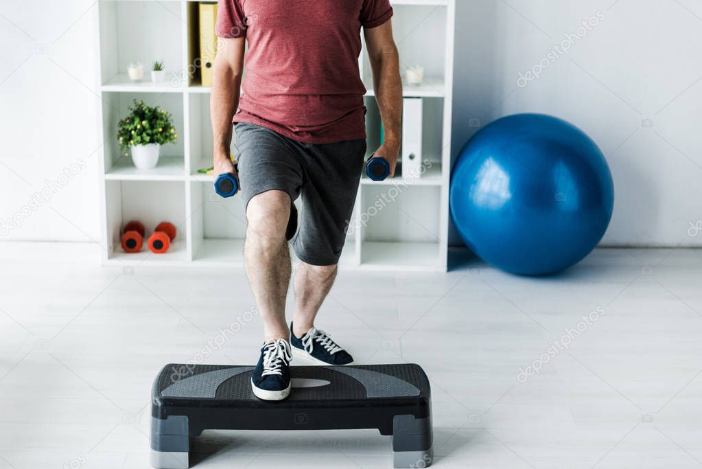 cropped view of middle aged man exercising with dumbbells on step platform in clinic  