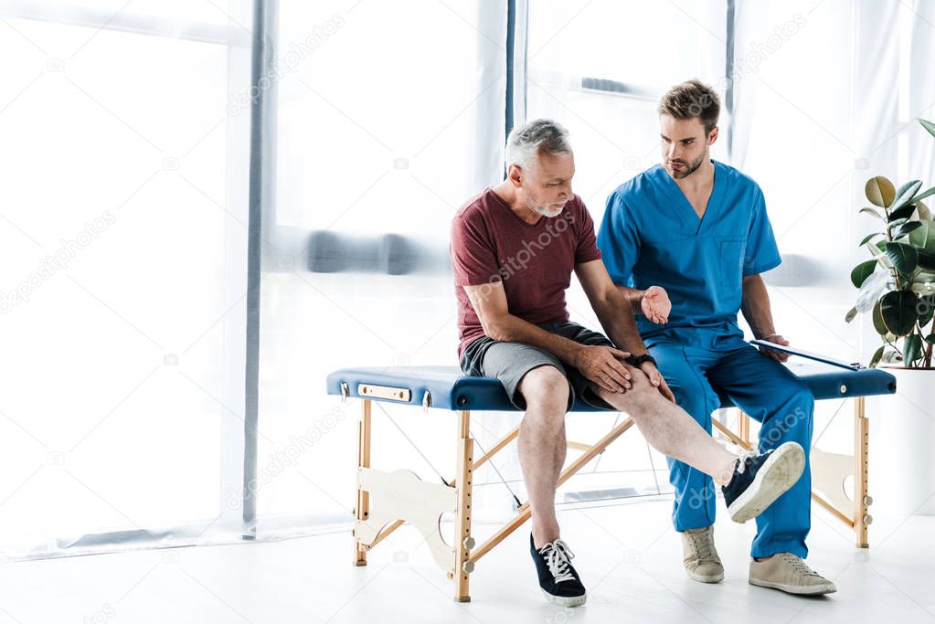 bearded mature patient touching leg near doctor in clinic 