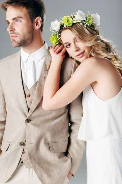 ウェディングドレスの花嫁と花輪は灰色に隔離されたスーツでハンサムな花婿を抱きしめる — ストック写真