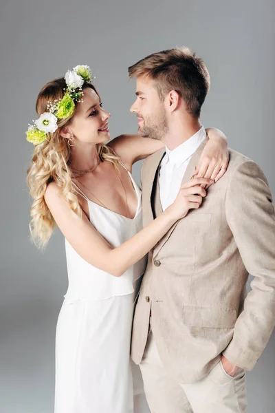 ウェディングドレスで花嫁と抱き合うスーツのハンサムな花婿とグレーに隔離された花輪 — ストック写真