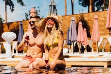 sexy couple holding glasses of red wine and smiling in swimming pool