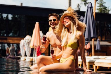 sexy couple holding glasses of red wine and smiling in swimming pool