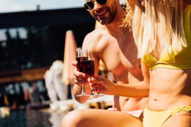 cropped view of sexy couple holding glasses of red wine in swimming pool