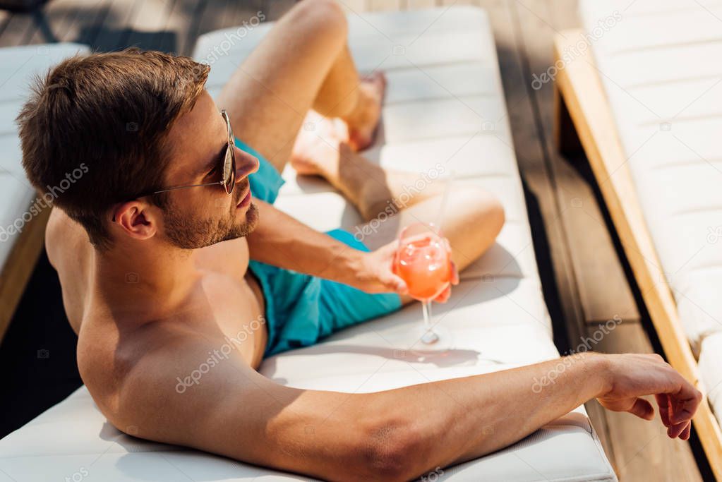 happy shirtless man in sunglasses lying on lounger and holding glass of cocktail at resort