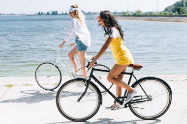 yaz aylarında nehir yakınında bisiklet sürme gülümseyen sarışın ve esmer kızların yan görünümü