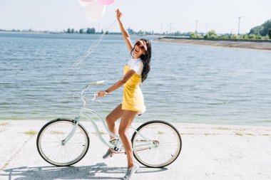 side view of happy brunette girl riding bike with balloons near river in summer clipart