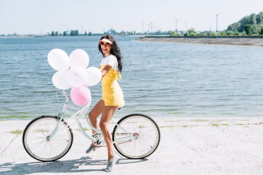 side view of beautiful brunette girl riding bike with balloons near river clipart