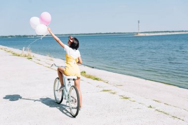 back view of brunette girl riding bike with balloons near river clipart