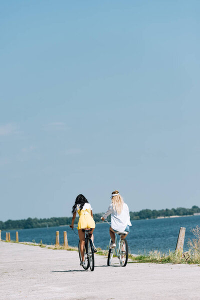 back view of blonde and brunette friends riding bikes near river in summer