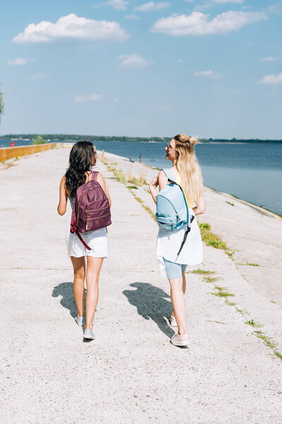 back view of girls walking with backpacks near river in summer