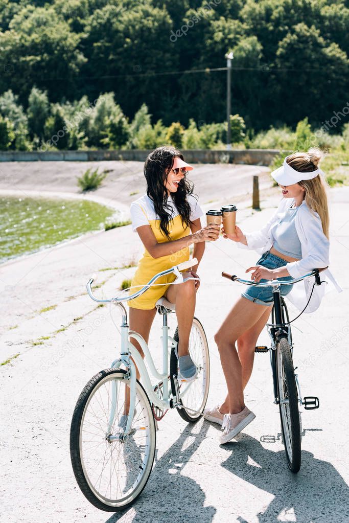 happy blonde and brunette girls with bikes and coffee to go near river in summer
