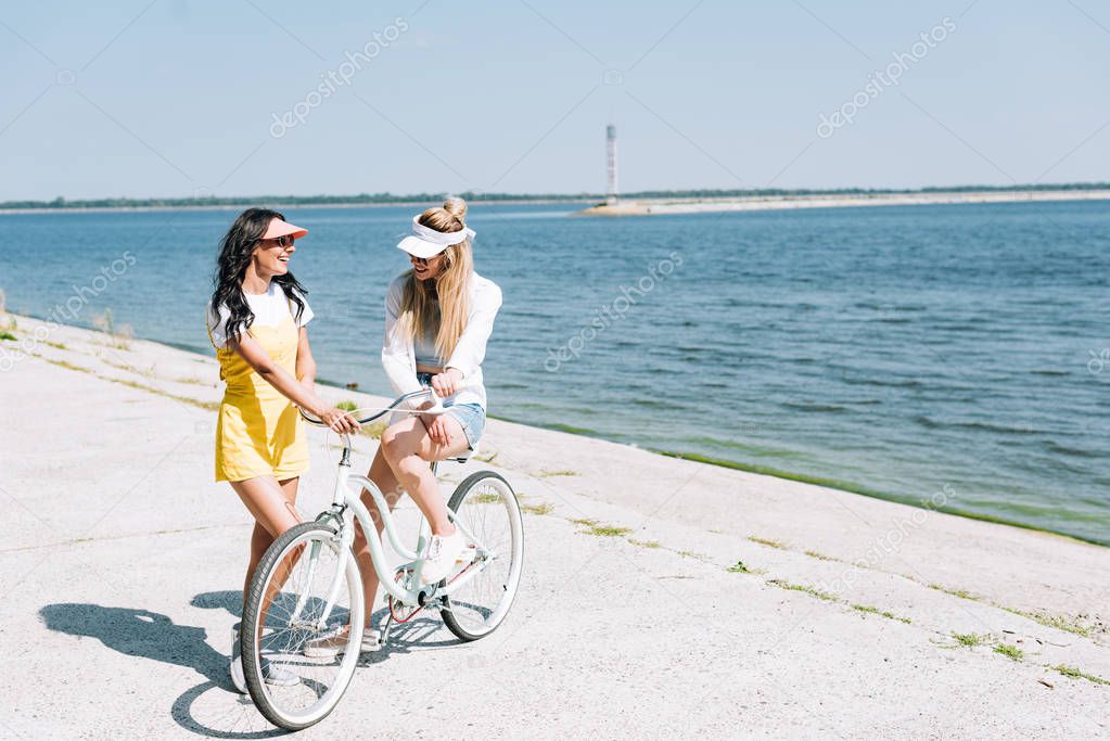 cheerful blonde and brunette girls with bike near river in summer