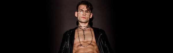 panoramic shot of brutal young man in glasses and leather jacket on black 